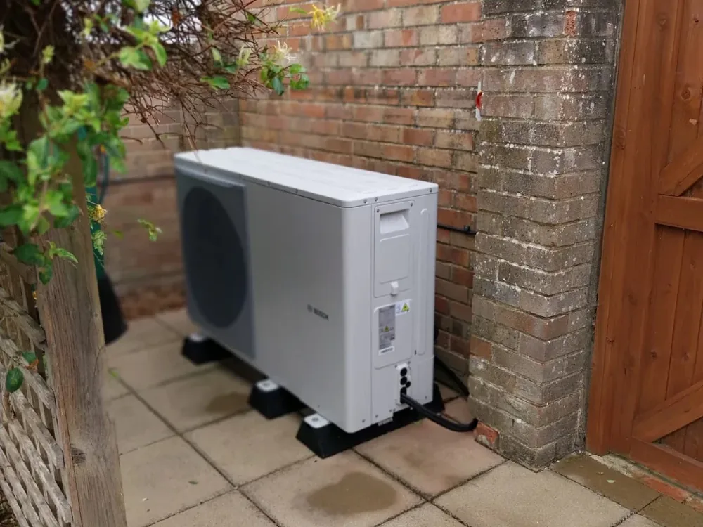 Outdoor air source heat pump unit standing on feet on a paved area beside the brick wall of a house, next to a timber gate, with pipework running into the wall.